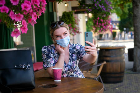 Beautiful young woman in a cafe in a medical mask and makes a selfie on the phone. A girl in a cafe on the street communicates online on a smartphone. Pandemic , quarantine.の写真素材