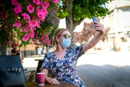 Beautiful young woman in a cafe in a sunglasses and medical mask and makes a selfie on the phone. A girl in a cafe on the street communicates online on a smartphone. Pandemic , quarantine.の写真素材
