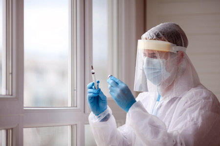 A doctor or healthcare professional in a personal protective kit, wearing rubber gloves in a hospital room in an epidemic or pandemic of coronavirus or COVID 19, draws a syringe with medicine. Vaccineの写真素材