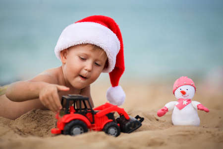 Happy boy in a santa claus hat plays with a toy car on the beach. A child in Christmas is resting on the sea. Copy spaceの写真素材