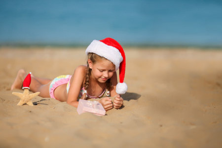 The child sits on the sand wearing a Santa hat. Little girl smiles and looks at a box with a gift. Merry Christmas. Winter vacation. Sea in the background. Copy space.の写真素材