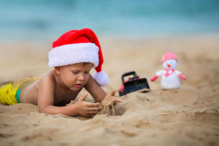 Happy boy in santa claus hat plays in the sand on the beach. A child in Christmas is resting on the sea. Copy spaceの写真素材