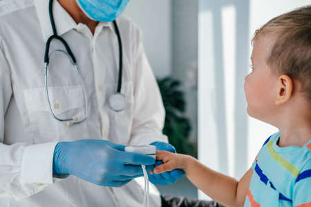 A doctor wearing medical gloves and a mask puts a pulse oximeter on the child's finger to measure the oxygen saturation. Little boy checks the oxygen in the blood in the doctor's office.の写真素材