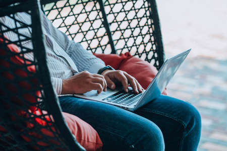 A man is sitting on a hanging rope swing and working on a laptop. Male hands on the keyboard are shopping online. Online shopping, online work.の写真素材