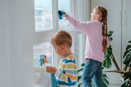 Little boy and girl sprinkles water from a bottle on the window and wipes it off with a rag. The children helps with cleaning the house. Clean the window. Brother and sister busy cleaning.の写真素材