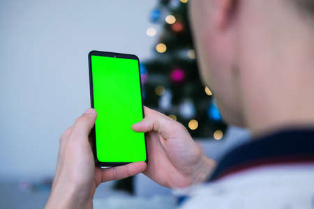 Closeup shot of man hand using mobile phone with Green screen against the backdrop of a Christmas tree. Finger swiping up and down in green screen of the mobile. Green screen mockup template.の写真素材