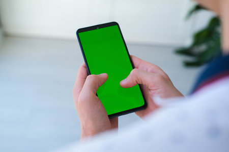 Man holds mobile phone and playing games online indoors of cozy home. Use green screen for copy space close up. Chroma key mock-up on smartphone in hand.の写真素材