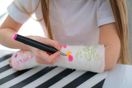 A little girl paints a cast on her arm. A child draws a drawing on a cast after a hand injury.の写真素材