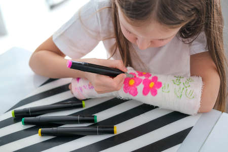 A little girl paints a cast on her arm. A child draws a drawing on a cast after a hand injury.の写真素材