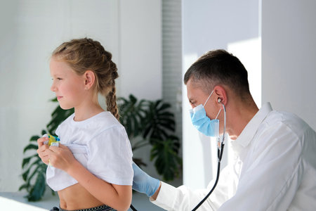 A doctor in a medical mask listens to the lungs of a child with a stethoscope. A doctor examines a little girl during an illness. Calling a doctor at home.の写真素材