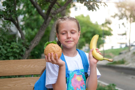 A schoolgirl girl sits on a bench and cannot choose what to eat. Choice of healthy or unhealthy food. Child chooses between burger and bananaの写真素材