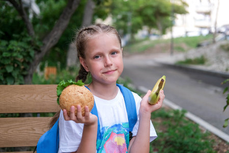 A schoolgirl girl sits on a bench and cannot choose what to eat. Choice of healthy or unhealthy food. Child chooses between burger and bananaの写真素材