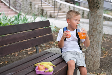 A little boy sits on a bench and drinks juice. A child with a backpack snacks on fruit at recess at school.の写真素材