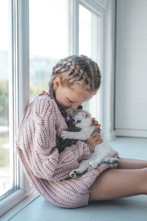 A cute little girl in a knitted sweater holds a kitten in her arms. The child plays with the cat and laughs.の写真素材