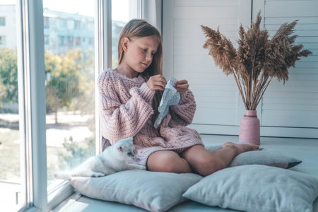 The child sews a soft toy near the window and plays with the kitten. The girl sews up a soft toy of an elephant. handmade, hobby.の写真素材