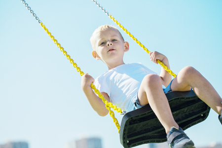 Little boy looks into the camera and enjoys the swings on the playground in the city park on a sunny summer day, close-upの写真素材