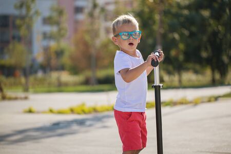 Handsome little boy in stylish sunglasses rides a scooter on a track in the park on a sunny summer dayの写真素材
