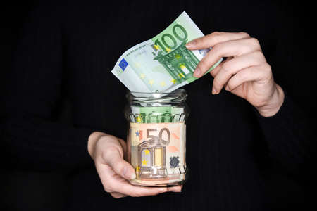 Woman holding a jar full of money. Woman pulling money out of the jar. Saving money in a jar.の写真素材