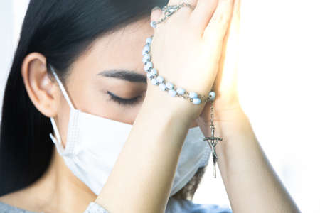Coronavirus outbreak: A woman wearing a medical mask praying over the coronavirus global pandemic to reduce anxiety and depression. Woman holding rosary praying.の写真素材