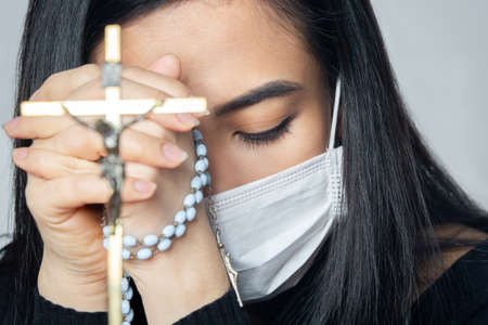 Coronavirus outbreak: A woman wearing a medical mask praying over the coronavirus global pandemic to reduce anxiety and depression. Woman holding rosary praying.