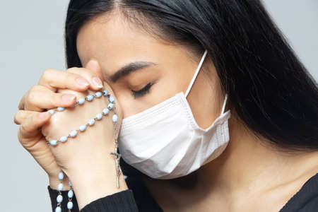 Coronavirus outbreak: A woman wearing a medical mask praying over the coronavirus global pandemic to reduce anxiety and depression. Woman holding rosary praying.の写真素材