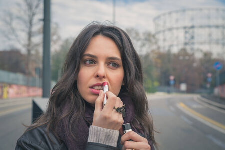 Beautiful young brunette with long hair putting on lipstick in the city streetsの写真素材