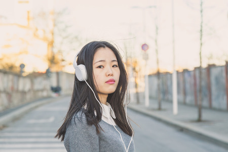Young beautiful Chinese girl with headphones listening to music in the city streetsの写真素材