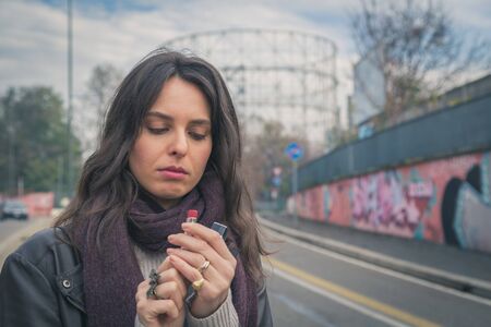 Beautiful young brunette with long hair putting on lipstick in the city streetsの写真素材