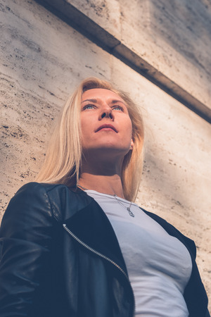 Beautiful blonde girl wearing ripped jeans and leather jacket posing in the city streetsの写真素材