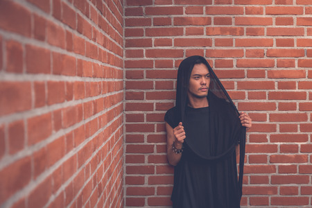 young handsome asian model dressed in black tunic posing with a brick wallの写真素材