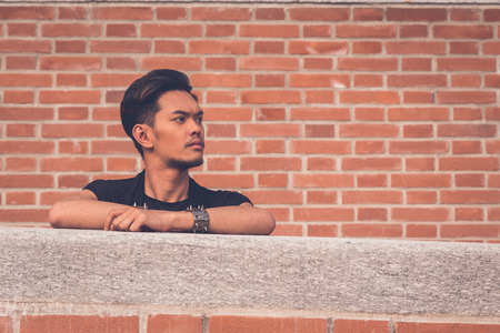 Young handsome Asian model dressed in black tunic posing with a brick wall in backgroundの写真素材
