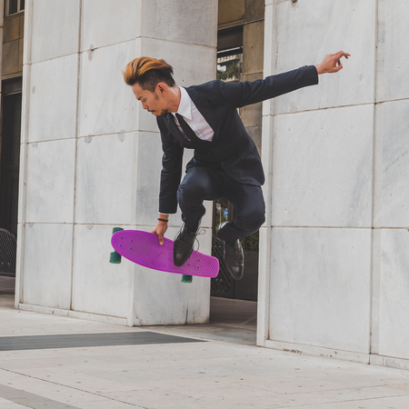 Young handsome Asian model dressed in dark suit and tie jumping with his skateboardの写真素材