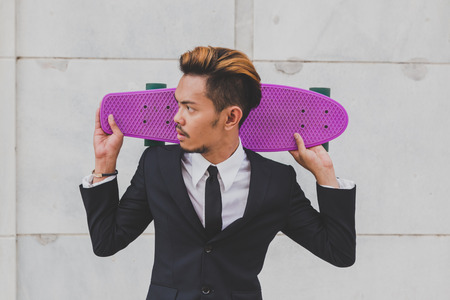 Young handsome Asian model dressed in dark suit and tie posing with his skateboardの写真素材