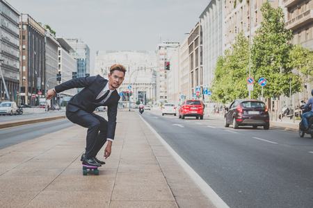 Young handsome Asian model dressed in dark suit and tie riding his skateboardの写真素材
