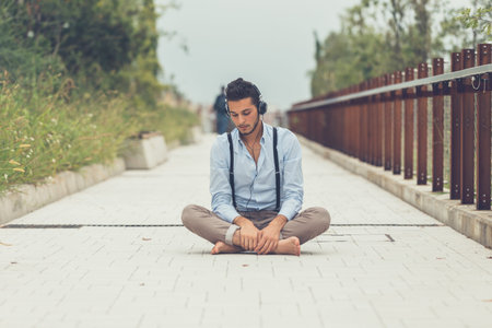 Young handsome man with short hair and beard wearing suspenders and listening to music in an urban...