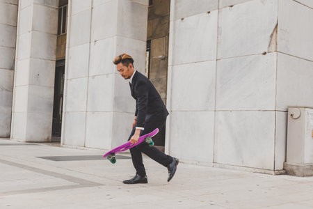 Young handsome Asian model dressed in dark suit and tie posing with his skateboard