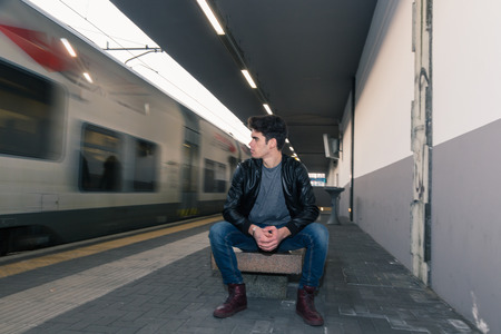 Beautiful young man with short hair sitting on a bench in a train stationの写真素材