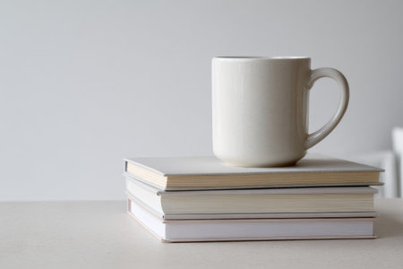 A stack of books supporting a white mug against a neutral background on a light-toned surfaceの素材