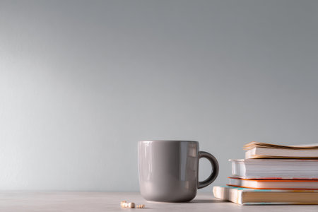 A gray coffee mug sits beside a stack of books with pills scattered on a light surfaceの素材
