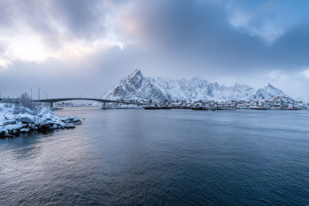 A snow-covered bridge spans dark water, leading to a mountain range and townの素材