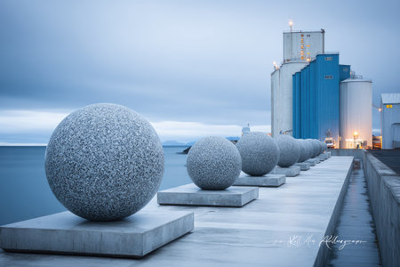 Series of stone spheres on plinths overlook calm water near industrial structures at duskの素材