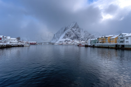 Scenic winter harbor town framed by snow-covered mountains under a cloudy skyの素材
