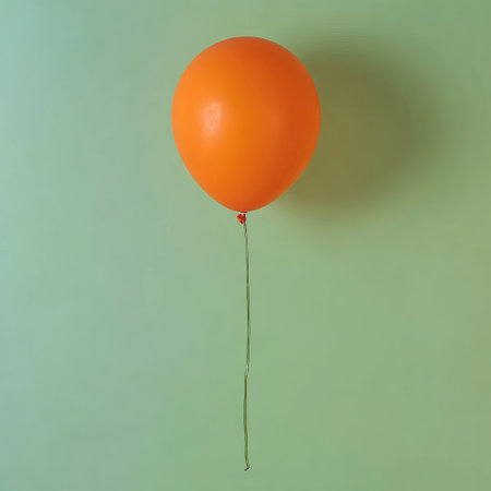 A bright orange balloon, tethered to a string, floats against a soft green backdropの素材
