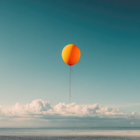 A single, bright orange balloon floats in the vast blue sky above a cloud bankの素材