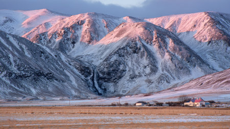 The image features snow-covered mountain peaks under a vibrant sunrise. The composition showcases the rugged terrain, with hues of pink and orange illuminating the snow. A small building sits in the valley below, under a clear sky, hinting at a remote setting. Suitable for various editorial and commercial applications.の素材