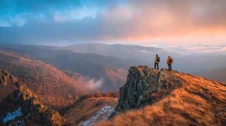 Two people stand atop a mountain ridge, gazing at a panoramic view during a vibrant sunrise or sunset. The scene displays a colorful sky and mountain range, with warm and cool tones. The photograph likely suits various commercial and editorial purposes, including travel and adventure themes.の素材