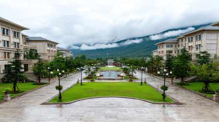 An aerial perspective captures a wide avenue flanked by symmetrical buildings and manicured lawns. The buildings feature light-colored facades, complemented by green trees. The composition includes a central pathway leading towards distant mountains shrouded in mist under an overcast sky, suitable for editorial and commercial use.の素材