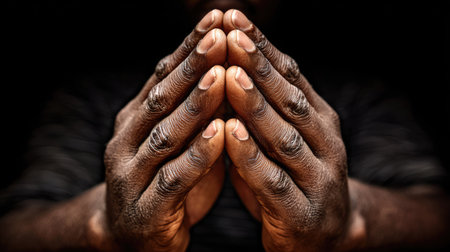The image captures a pair of hands clasped together, suggesting prayer or contemplation. The skin tone is dark, illuminated by selective lighting that highlights the texture and form of the hands. The backdrop is dark, emphasizing the subject. This composition could be used for various projects representing faith, reflection, or spirituality.の素材