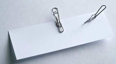 A close-up view displays a white rectangular paper with two metallic clips. The composition features clean lines and high-key lighting, enhancing the texture of the paper. This arrangement is suitable for presentations, advertising, or design projects. The minimalist approach offers copy space and versatile application.の素材