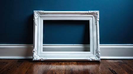 An empty white ornate picture frame leans against a dark blue wall. The composition is simple, with the frame's intricate details contrasting with the smooth wall. Wooden flooring adds texture and depth to the setting. The image could be suitable for visual design, showcasing art, or general interior design.の素材